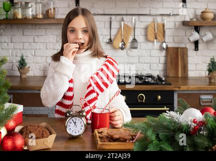 Das Kind isst Schokoladenkekse und hält einen Becher mit heißem Kakao in der Hand und blickt auf die Kamera in der weihnachtlich dekorierten Küche Stockfoto