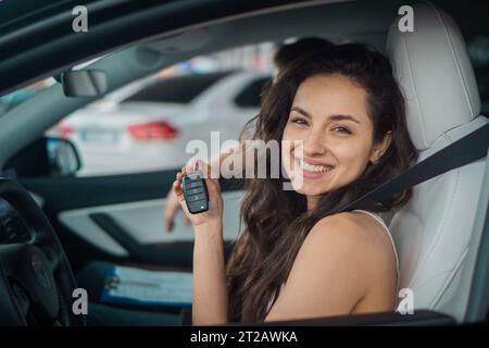 Glückliche, schöne Frau, die in einem modernen Auto sitzt und den Schlüssel für die Kamera zeigt. Mann auf Beifahrersitz. Reise, Lifestyle, Verkehrskonzept Stockfoto