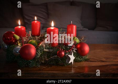 Adventskranz mit drei brennenden roten Kerzen und Weihnachtsdekoration auf einem Holztisch vor der Couch, festliche Wohnkultur für den dritten Sonntag Stockfoto