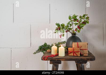 Natürliche Advents- und Weihnachtsdekoration, Geschenke, Kerzen und Zweige mit roten Früchten in einer Vase auf einem rustikalen Holzhocker vor einer rauen weißen Wand Stockfoto