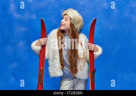 Winterfreude, fröhliches junges Mädchen in Kunstpelzjacke und Hut mit roten Skiern auf türkisfarbenem Hintergrund Stockfoto