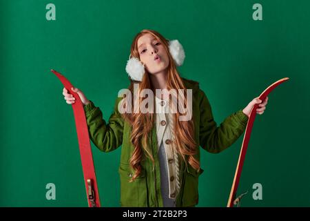 Kalte Luft, ein junges Kind in Ohrenschützern und ein Winteroutfit, das atmet und rote Skier auf Türkis hält Stockfoto