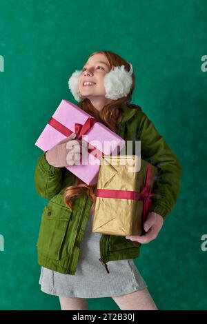 Saison der Freude, fröhliches Mädchen im Winteroutfit und Ohrenschützer, die Weihnachtsgeschenke unter fallendem Schnee halten Stockfoto