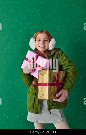 Saison der Freude, glückliches Mädchen im Winteroutfit und Ohrenschützer, die Weihnachtsgeschenke unter fallendem Schnee halten Stockfoto