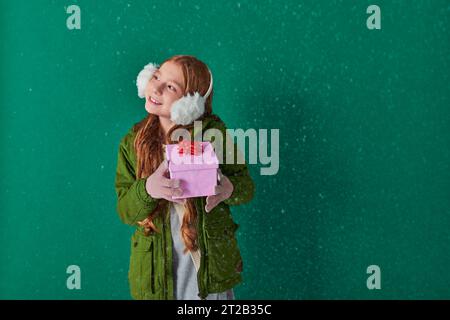 Saison der Freude, fröhliches Kind in Ohrenschützern und Winteroutfit mit Weihnachtsgeschenk unter fallendem Schnee Stockfoto