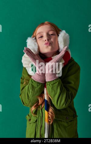 Ein junges Mädchen in Ohrenschützern, gestreiftem Schal und Winteroutfit mit türkisfarbenen Lippen und Luftküssen Stockfoto