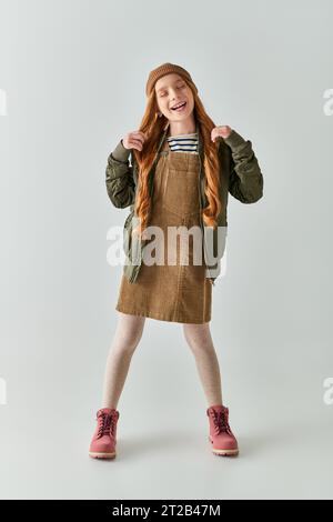 Zufriedenes Mädchen mit langen Haaren und Hut auf dem Kopf stehend im Kleid mit Winterjacke auf grauem Hintergrund Stockfoto