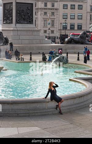 Eine attraktive junge weiße Europäerin in einem schwarzen Kleid posiert für Fotos an einem der Springbrunnen am Trafalgar Square, Central London England, Großbritannien Stockfoto