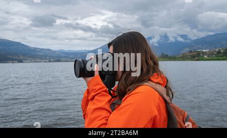 Nahaufnahme einer wunderschönen jungen lateinamerikanischen Frau in orangefarbener Jacke mit Rucksack, von der Seite gesehen, die Fotos neben einer Seenuunde macht Stockfoto