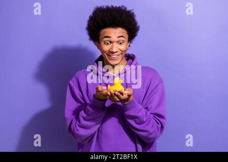 Porträt der beeindruckten positiven überfreudeten Person tragen violetten Hoodie Blick auf Hände halten kleines Entenspielzeug isoliert auf lila Hintergrund Stockfoto