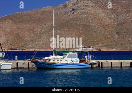 Blaues Segelboot, das im Hafen von Livadia mit den Bergen der hübschen Insel Tilos im Hintergrund vor Anker liegt. Dodekanese, Griechenland. Mai/Juni 2023 Stockfoto