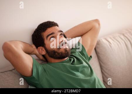 Nachdenklicher junger arabischer Mann mit Bart sitzt auf Sofa, ruht sich aus, schaut auf freien Raum, genießt Freizeit im Wohnzimmerinnenraum Stockfoto