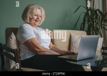 Inhalt ältere Frau mit kurzen Haaren, die ein weißes T-Shirt trägt und mit Laptop auf der Couch sitzt. Nutzung elektronischer Geräte, Videoanrufe von erwachsenen Kindern Stockfoto