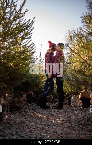 Wunderschönes Paar in karierten roten Hemden, Strickmützen umarmen sich von Angesicht zu Angesicht zwischen grünem Weihnachtsbaummarkt. Junger Mann lacht Stockfoto