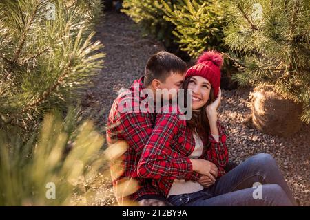 Nahaufnahmen von jungen Paaren in roten karierten Hemden, die auf dem Straßenmarkt zwischen Weihnachtsbaumkeimlingen sitzen. Mann umarmen Frau in Strickmütze W Stockfoto