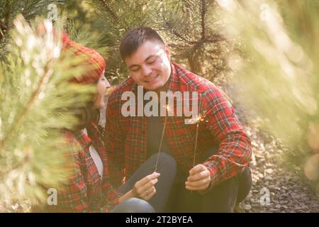 Durch grüne Nadelbäume aus Kiefern halten junge glückliche Paare in roten karierten Hemden festliche Funkeln im Wald. Mann und Frau mit Hut lachen über Christus Stockfoto