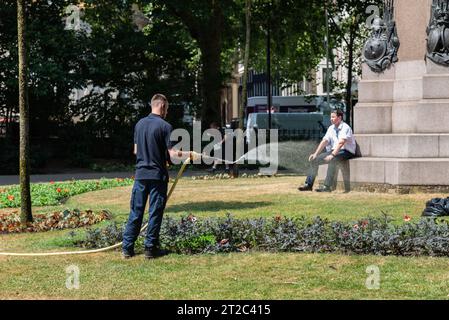 Sehr heißer Tag in London, Großbritannien. Ein Gärtner, der die Pflanzen in Victoria Embankment Gardens in der Nähe eines vermeintlichen Büroarbeiters in der heißen Hitze bewässert Stockfoto