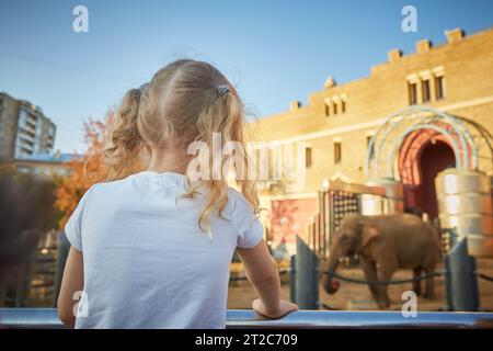 Ein gesichtsloses Mädchen mit lockigen Haaren schaut begeistert einen Elefanten im Zoo an. Das Konzept der Erholung und Unterhaltung. Stockfoto