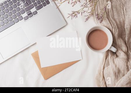 Kaffeetasse, Laptop, Umschlag mit leerer Karte und kleinen Blumen auf weißer Bettwäsche. Gemütlicher Arbeitsplatz, Frühstück, Komfort, Hygge-Konzept. Draufsicht, f Stockfoto