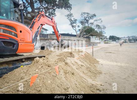 Bulldozer gräbt Sand an einem Sandstrand. Nicht erkennbare Leute, die am Strand spazieren gehen. Auckland. Stockfoto