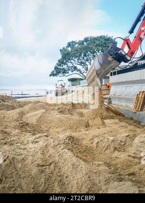 Bulldozer gräbt Sand an einem Sandstrand. Häuser und Betonmauern am Strand. Vertikales Format. Stockfoto