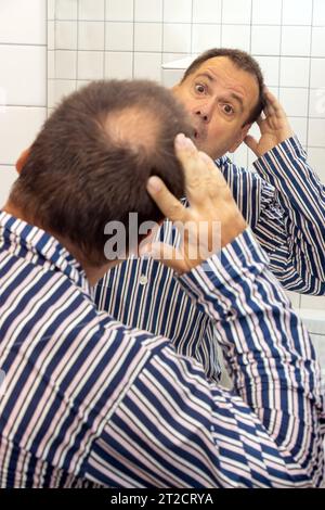 Ein überraschter Mann im Pyjama beobachtet den Beginn der Kahlheit auf seinem Kopf im Spiegel Stockfoto