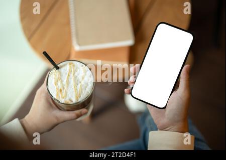 Nahaufnahme einer Frau, die ein eiskaltes Kaffeeglas und ein Smartphone-Modell mit weißem Bildschirm hält, während sie in einem Café sitzt. Technologie und Lifestyle c Stockfoto