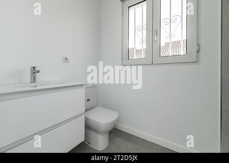 Kleines Badezimmer in einem Haus mit weißem Porzellanwaschbecken an der Wand mit Holzschubladen und weißem Aluminiumfenster Stockfoto