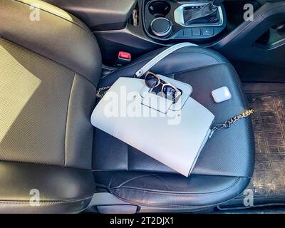 Die wunderschöne, modische Ledertasche und Sonnenbrille mit kabellosen Kopfhörern für weiße Frauen liegen auf dem Ledersessel eines teuren Autos. Stockfoto