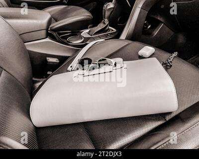 Die wunderschöne, modische Ledertasche und Sonnenbrille mit kabellosen Kopfhörern für weiße Frauen liegen auf dem Ledersessel eines teuren Autos. Stockfoto