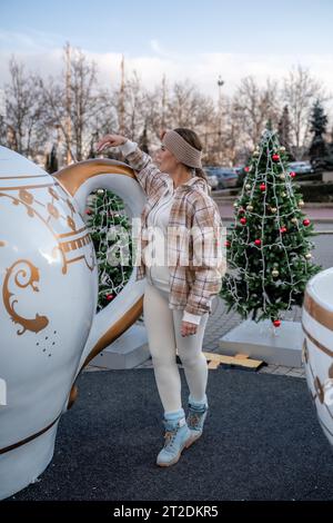 Frauenweihnachtsplatz. Sie steht in der Nähe einer großen weißen Tasse, in einem leichten Anzug gekleidet. Mit Bäumen, die mit weihnachtlichem Lametta im Hintergrund verziert sind Stockfoto