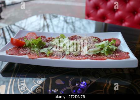 Fleischvorspeise aus rotem Rindfleisch und Schweinefleisch, bestreut mit Käse und Gemüse auf einem weißen rechteckigen Teller auf einem Tisch in einem Café, Restaurant. Stockfoto