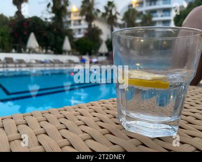 Hausgemachte Limonade in Gläsern mit Zitronenscheibe, Zitronenkorb, Trinkhalm, auf weißem Tisch, am Rand des Pools mit türkisblauem Wasserhinterm Stockfoto