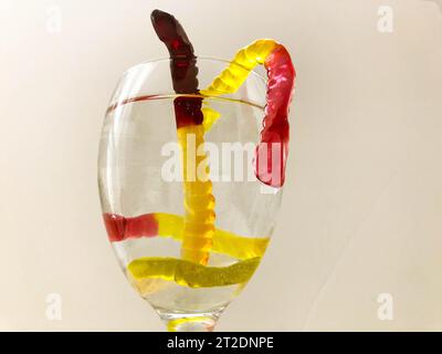 Gummiwürmer schwimmen in einem transparenten Glas Wasser. Helle, süße Gummiwürmer klettern aus dem Wasser. Gelbe, braune, rote Gelatinewürmer im Glas Stockfoto