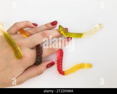 Lange, mundtränende, mehrfarbige Würmer liegen mit einer knallroten Maniküre auf der Hand des Mädchens. Grüne, gelbe, braune und transparente Zuckerwürmer kriechen alle Stockfoto