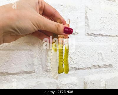 Ein Mädchen mit einer hellroten Maniküre hält gummiartige, helle Würmer vor dem Hintergrund einer weißen Ziegelwand. Süßigkeiten an einer verputzten Wand. Helles, gelbes Ma Stockfoto