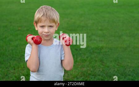 Sportlicher kleiner Junge, der Kurzhanteln auf einem grünen Rasen hebt. Fröhliches Kleinkind, das im Freien Sport vor dem Hintergrund der Natur macht. Happy Child hält roten Dumbbel Stockfoto