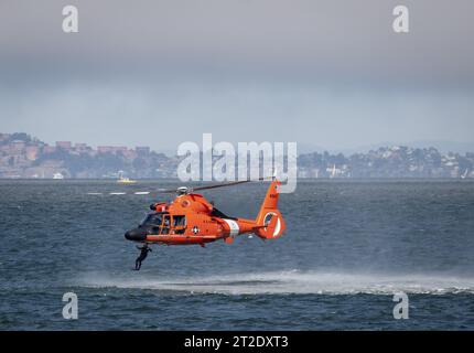 Aus der Vogelperspektive eines leuchtenden orangefarbenen Hubschraubers, der über ein riesiges blaues Gewässer fliegt, mit einer weitläufigen Stadtlandschaft im Hintergrund Stockfoto