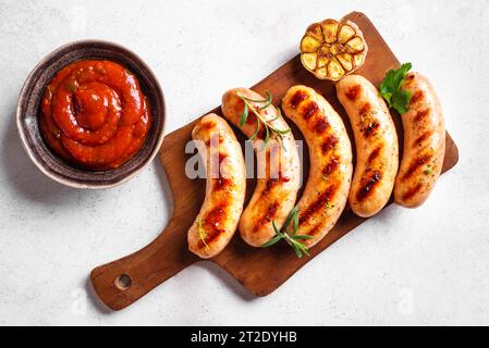Gegrillte Würstchen mit Tomatensauce und Kräutern auf weißem Hintergrund, Draufsicht, Kopierraum. Gebratene hausgemachte Würstchen auf Holzbrett. Stockfoto