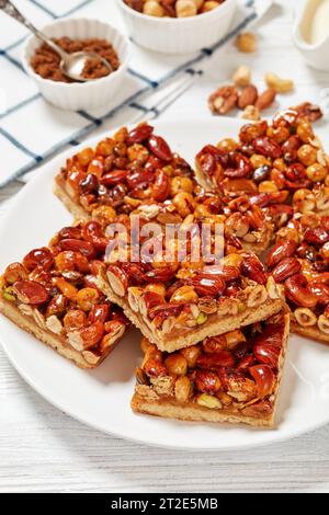 Rustikale Toffee gemischte Nüsse Shortbread Bars auf weißem Teller auf weißem Holztisch mit Zutaten im Hintergrund, vertikale Ansicht, Nahaufnahme Stockfoto