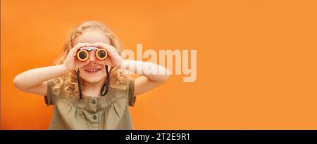 Ein süßes Mädchen mit lockigen Haaren schaut durch ein Fernglas auf einem monochromen Hintergrund im Studio. Kopierbereich Stockfoto