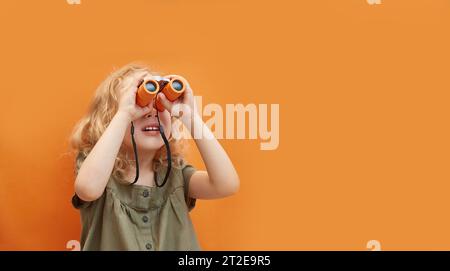 Ein süßes Mädchen mit lockigen Haaren schaut durch ein Fernglas auf einem monochromen Hintergrund im Studio. Kopierbereich. Das Konzept von Reisen und Erholung, V Stockfoto