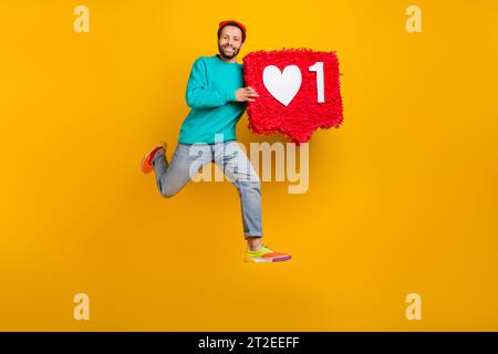 Foto in voller Größe von optimistischen Guy Wear Jeans rote Beanie Hut fliegender Lauf mit großem rotem Herz in den Händen isoliert auf gelbem Hintergrund Stockfoto