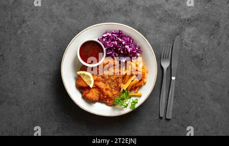 Schnitzel mit Kartoffelfrites, Rotkohlsalat und Sauce auf weißem Teller auf dunklem Steinhintergrund. Draufsicht, flach Stockfoto