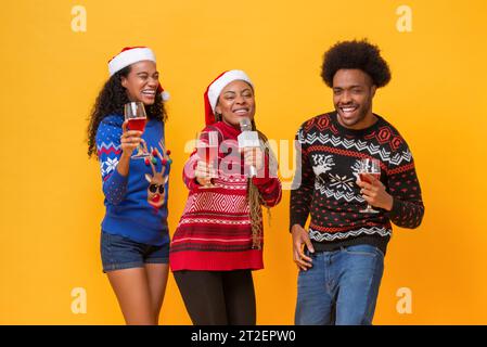 Glückliche, vielfältige Freunde in Weihnachtspullover trinken, singen, feiern und gemeinsam Spaß haben in gelbem isoliertem Hintergrundstudio Stockfoto