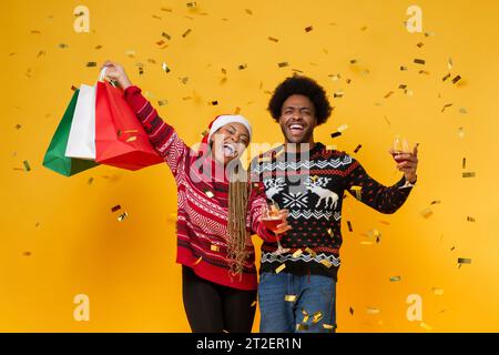 Afrikanisches Amercianpaar, das Pullover trägt, die Einkaufstaschen und Getränke halten, um Weihnachten im gelben Studiohintergrund zu feiern Stockfoto