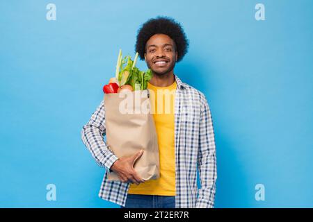 Gutaussehender freundlicher, lächelnder Afro-Afroamerikaner, der eine Einkaufstasche voller Lebensmittel in einem hellblauen isolierten Studio-Hintergrund hält Stockfoto