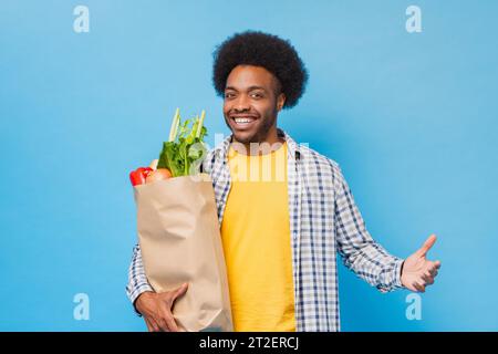 Gutaussehender freundlicher, lächelnder Afro-Afroamerikaner, der eine Einkaufstasche voller Lebensmittel in einem hellblauen isolierten Studio-Hintergrund hält Stockfoto