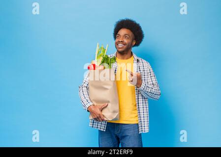 Hübscher, freundlicher, lächelnder Afro-Afroamerikaner, der eine Einkaufstasche voller Lebensmittel hält und Hand in hellblauem, isoliertem Studi kopiert Stockfoto