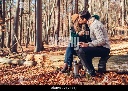 Glückliches, liebevolles Paar küsst Tee und entspannt im Herbstwald. Mann und Frau sitzen auf dem Kofferraum. Die Leute genießen das Date Stockfoto